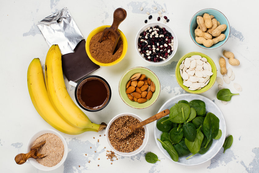 Imaginea arată alimente bogate în magneziu, cum ar fi banane, ciocolată neagră, spanac, migdale, semințe, boabe de fasole și cacao, aranjate pe o suprafață deschisă la culoare.